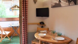Una habitación acogedora con una mesa de comedor de madera dispuesta para dos personas, un televisor montado en la pared y una ventana con cortinas.