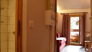 Un baño con ducha, inodoro y un perchero para toallas. En la habitación contigua, hay una cama con una manta roja y una silla de madera.