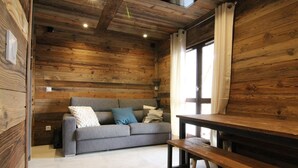 A cozy room with wooden walls, a grey sofa with cushions, a wooden bench, and a window with curtains. - Sunny 3-Room Apartment for 6 with Large Terrace, Parking & Ski-In/Ski-Out Access (Huez)