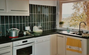 Uma cozinha com bancada preta, armários brancos, janela com vista para árvores e uma toalha com a inscrição 'ROKEMAR'.