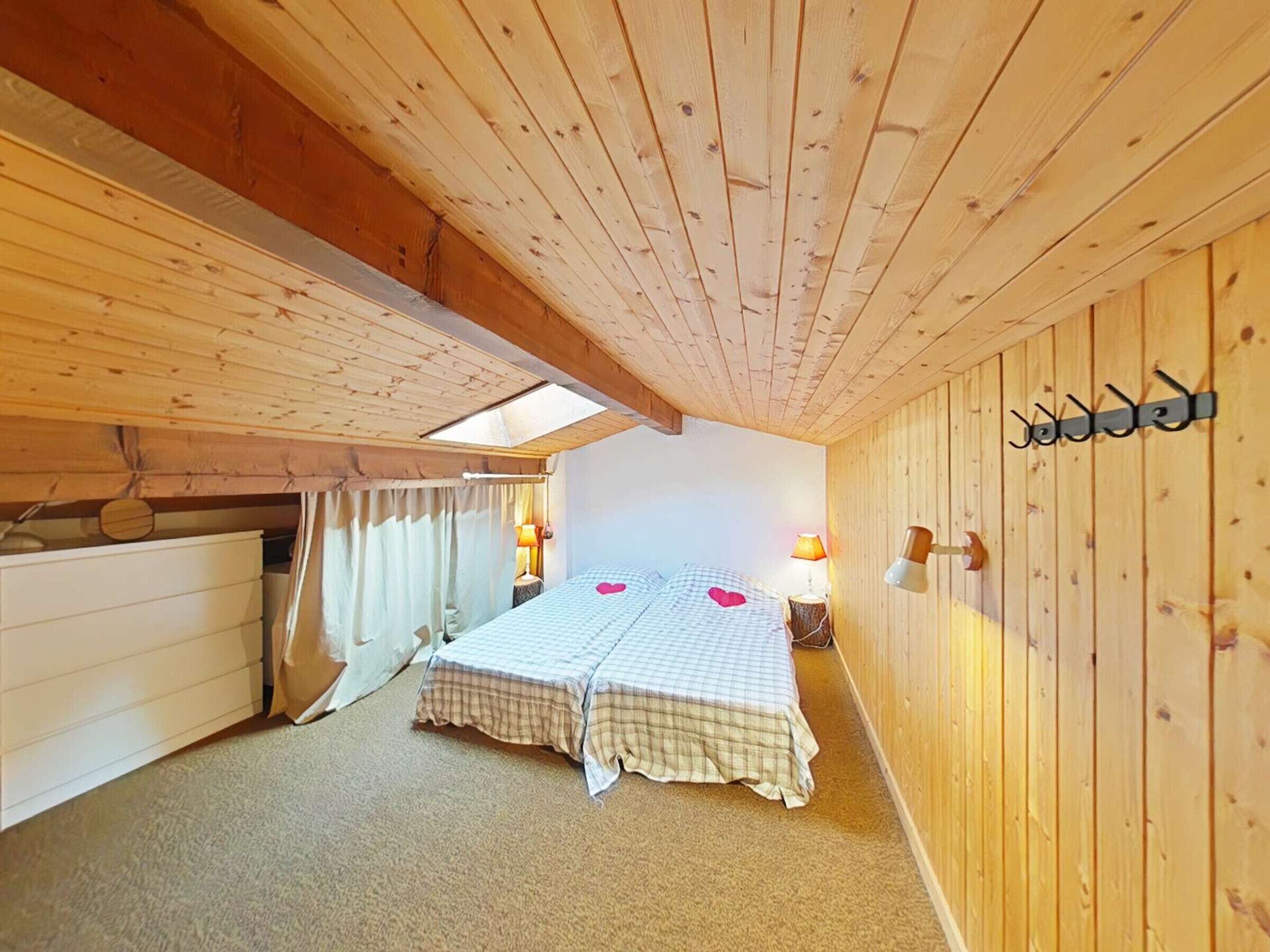 Une pièce chaleureuse aux murs et au plafond en bois, un lit recouvert d’une courtepointe à carreaux, une petite table avec une lampe et un porte-manteau fixé au mur.