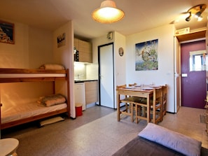 A compact room with a bunk bed, a small kitchenette, a dining table with chairs, and a door leading to another room.