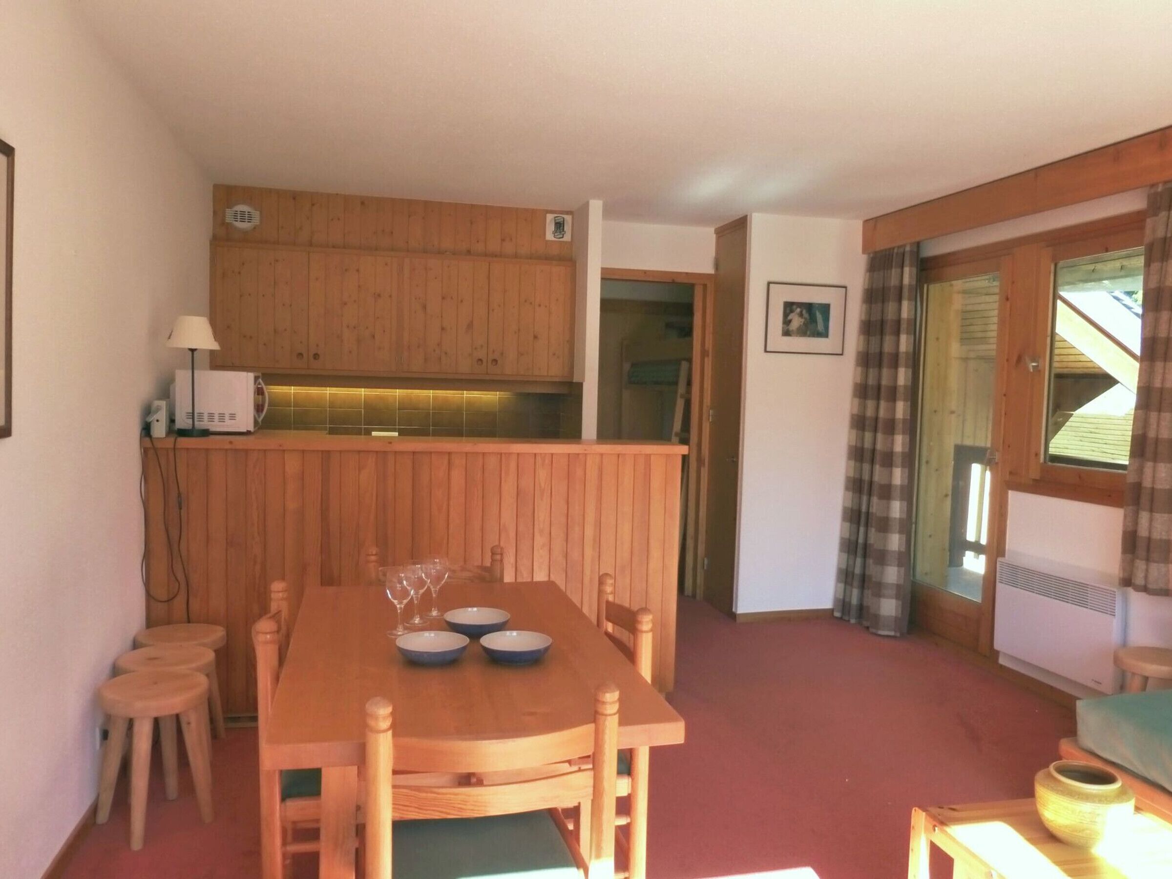 A cozy room with a wooden dining table set for four, a small kitchen area with wooden cabinets, and a window with curtains.