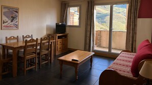 Una habitación con una mesa de comedor de madera, sillas, un televisor en un mueble bajo, un sofá y un amplio ventanal con cortinas.