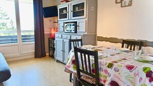 Comedor con una mesa dispuesta para cuatro, zona de cocina con refrigerador y armarios, y ventana con cortinas.