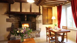 A cozy room with a stone fireplace, wooden beams, a dining table with chairs, and a window with red curtains. - Stone house in peace with pets allowed (Ouville)