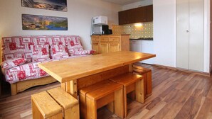 A compact living space with a wooden dining table, benches, a kitchenette, and a sofa with patterned cushions. - Cozy 2-Room Apartment for 6 in Alpe d'Huez, Ski-in/Ski-out, Near Shops, South-Facing Balcony (Huez)