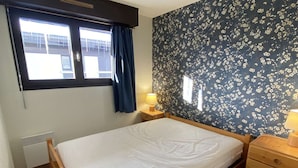 A bedroom with a bed, a nightstand, a dresser, a window with curtains, and a wall with a floral pattern. - Cozy 2-Room Apartment for 6 in Alpe d'Huez, Ski-in/Ski-out, Near Shops, South-Facing Balcony (Huez)