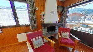 A cozy living room with wooden walls, a fireplace, two red armchairs, and a large window with a view of buildings and hills. - Studio Cabin 4 Sleeps, Ski-In/Ski-Out, South-Facing Balcony, Le Grand-Bornand (Le Grand-Bornand)
