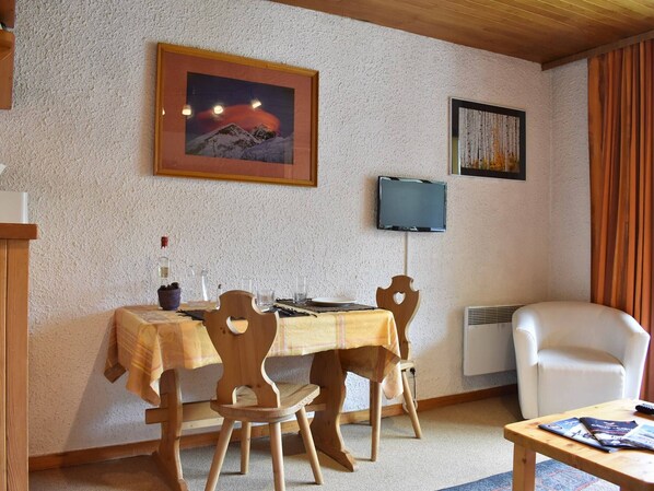 Une pièce chaleureuse avec une table à manger en bois, des chaises, un téléviseur fixé au mur et une fenêtre avec des rideaux.