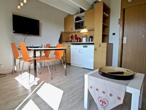 Eine kompakte Küche mit Mikrowelle, einem weißen Geschirrspüler und Holzausbau. Es gibt einen Esstisch mit orangefarbenen Stühlen und einen an der Wand montierten Fernseher.