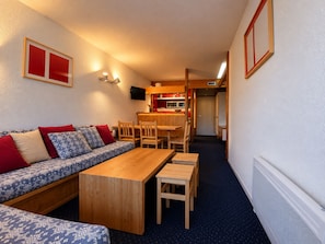 Un espace de vie compact comprenant un canapé, une table et des chaises en bois, un coin cuisine et une porte donnant sur une autre pièce.