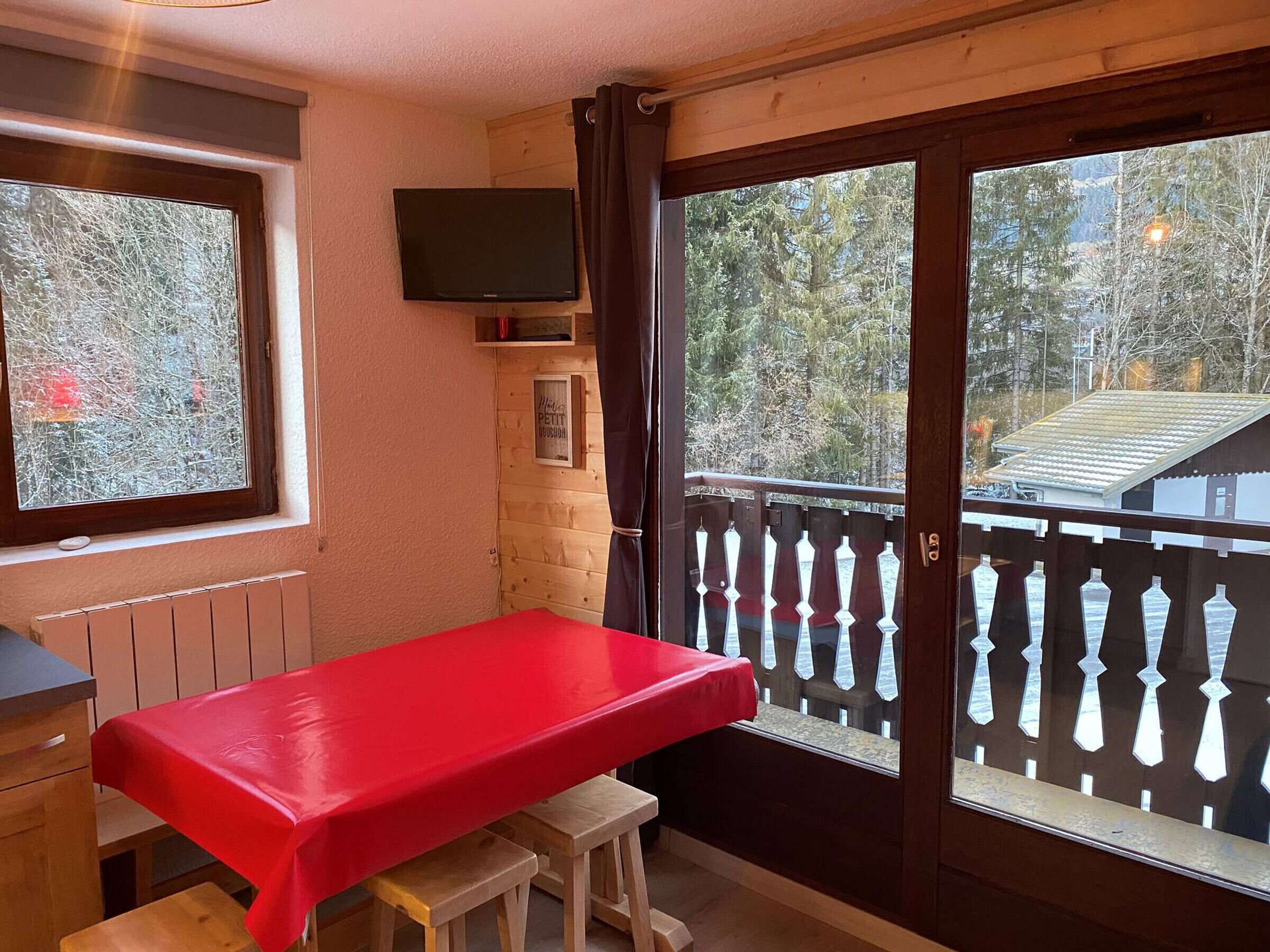 Un balcon avec une table rouge et des tabourets en bois, un téléviseur fixé au mur, et une vue sur des arbres et un immeuble à l’extérieur.