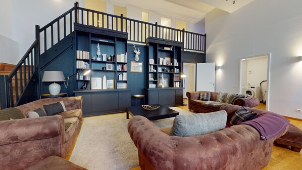 Amplia sala de estar con una escalera azul oscuro, sofás de cuero, una mesa de centro y una estantería llena de libros y objetos decorativos.