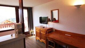 A hotel room with a wooden dining table, a TV mounted on the wall, a small wooden cabinet, and a balcony with a railing. - Cozy 2-Room Apartment for 6, Near Ski Lifts & Shops - L'Alpe d'Huez (Huez)