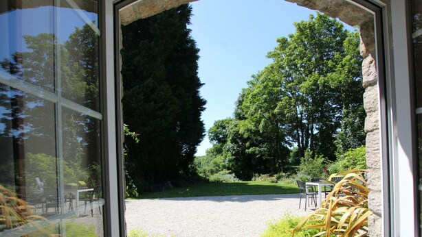 A view through a glass door into a garden with a gravel path, chairs, and greenery. - Cozy detached house with fireplace and enclosed garden in Normandy (Couville)