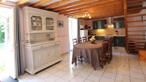 A dining area with a table and chairs, a white cabinet with glass doors, and a kitchen area with cabinets and appliances. - Cozy detached house with fireplace and enclosed garden in Normandy (Couville)