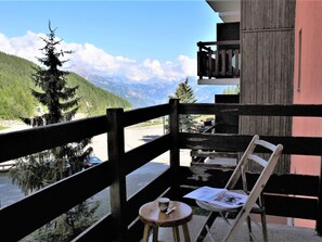 Ein Balkon mit Holzbalkon, ein kleiner Tisch mit einer Tasse und einer Zeitung, sowie ein Stuhl. Im Hintergrund ist eine Berglandschaft zu sehen.