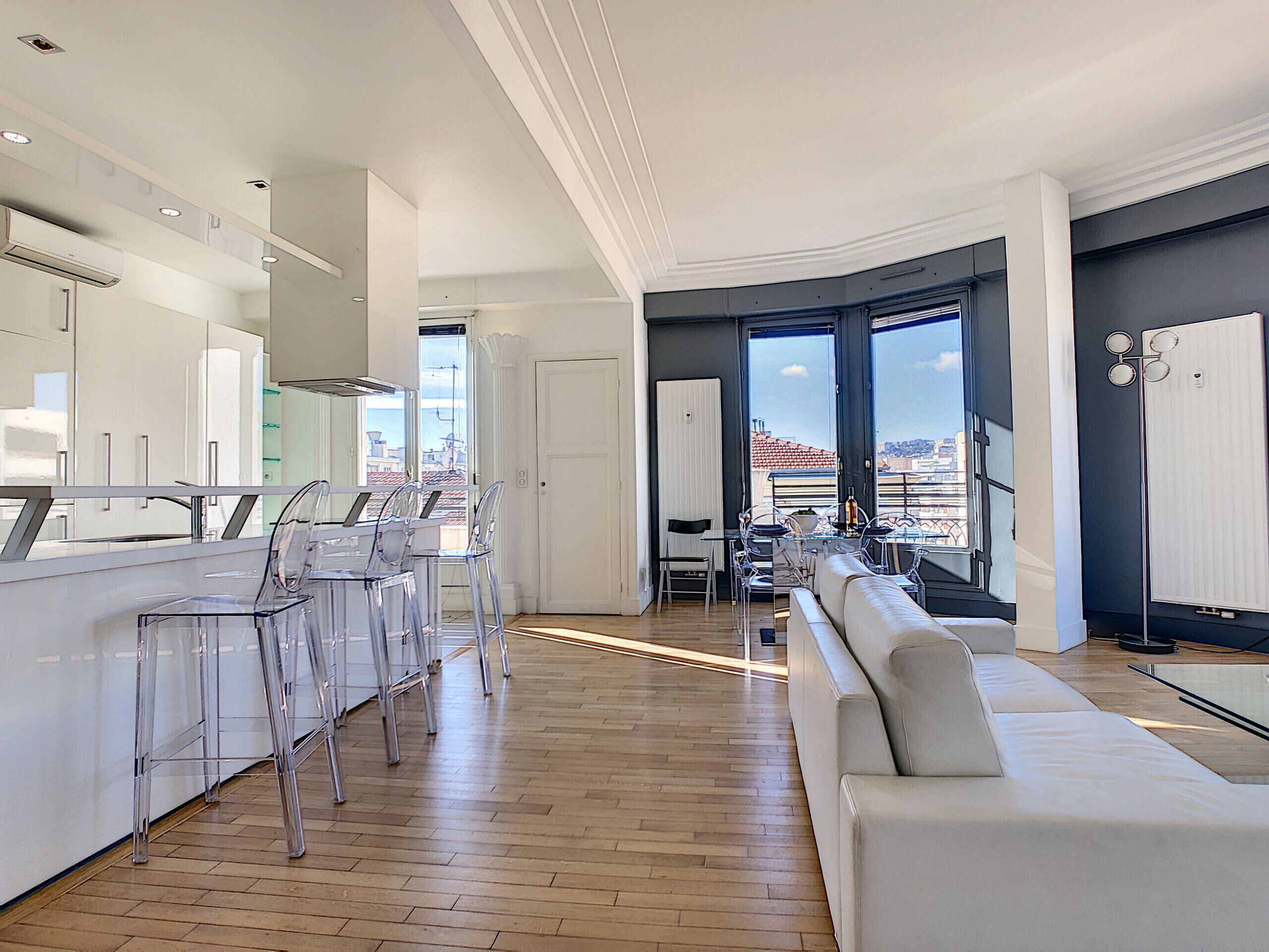 A modern kitchen with a central island, clear chairs, and a large window offering a city view.
