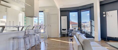 A modern kitchen with a central island, clear chairs, and a large window offering a city view.