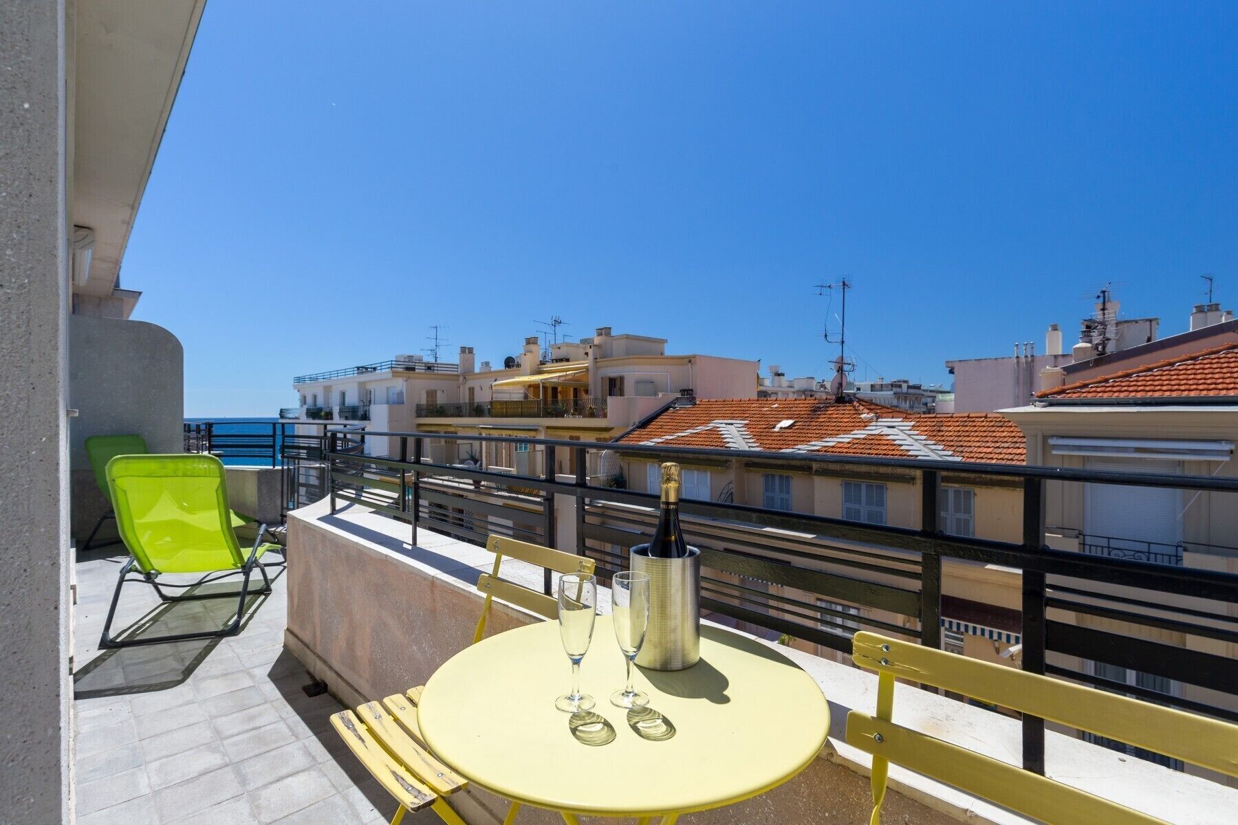 Ein Balkon mit einem gelben Tisch, zwei grünen Stühlen und einer Champagnerflasche.