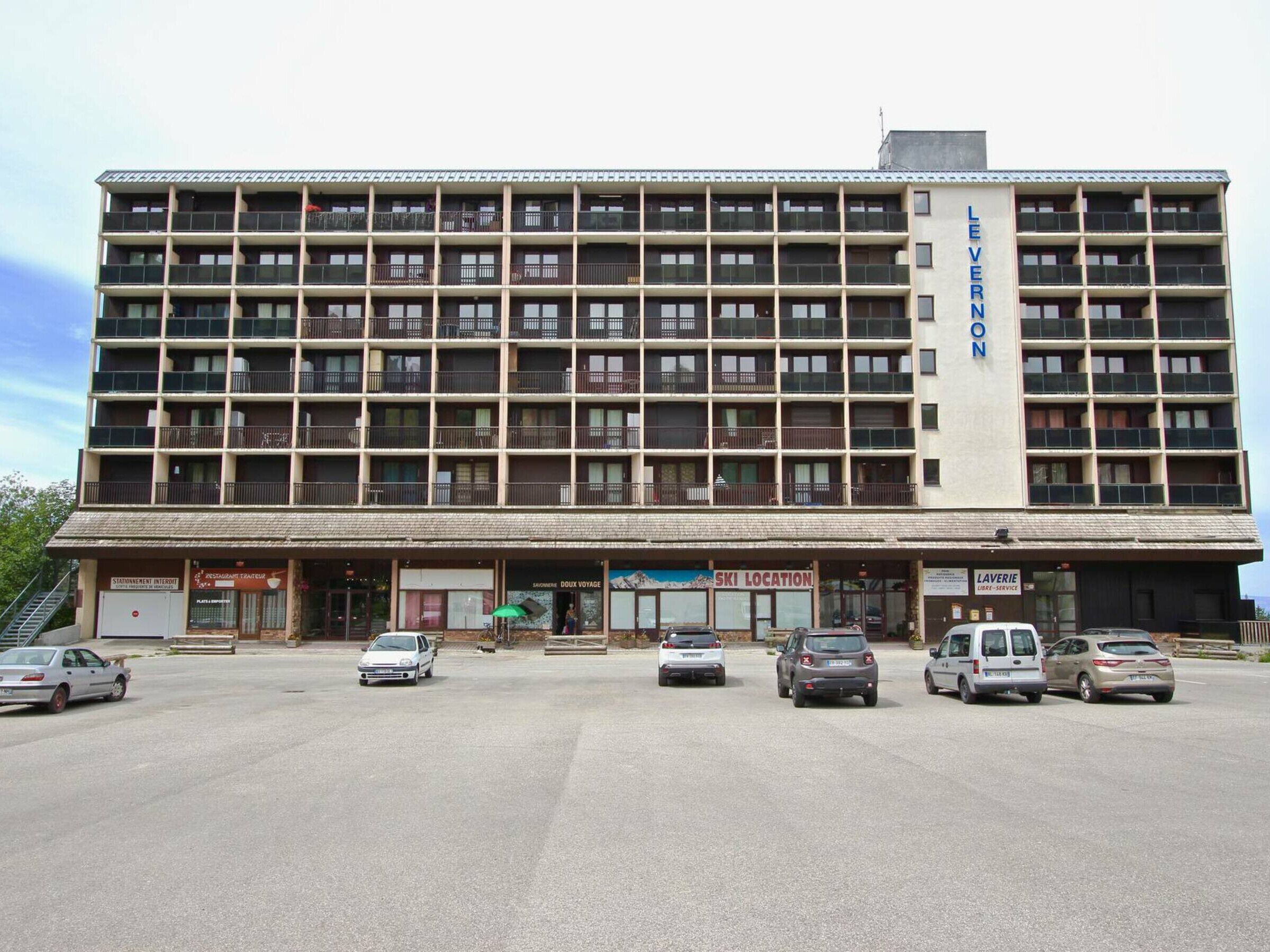 Un hotel a più piani con un ingresso centrale, diversi balconi e un parcheggio con auto.