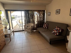 Sala de estar com sofá, uma cadeira, mesa pequena e porta de vidro deslizante que dá para área externa.