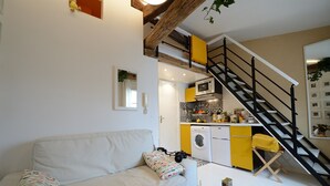 A compact living space with a sofa, a washing machine, a microwave, and a staircase leading to an upper level. - E5DG Discovery (París)