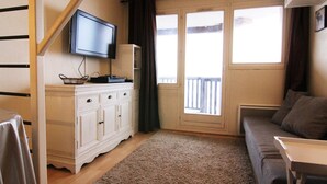 A living room with a flat-screen TV mounted on a white cabinet, a grey sofa, a small wooden table, and a large window with a balcony view.