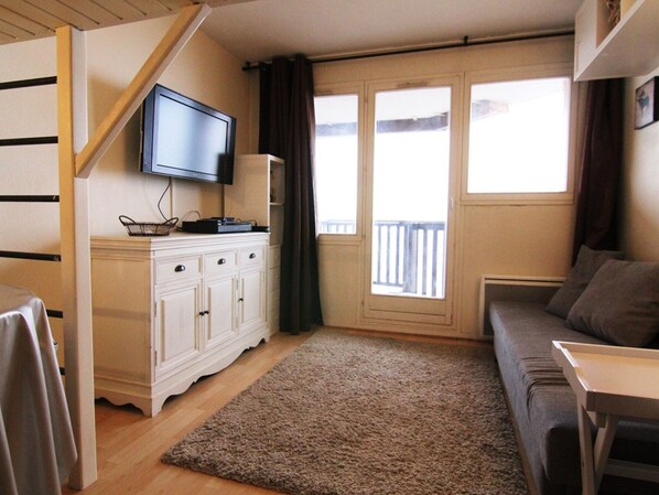 Una sala de estar con un televisor de pantalla plana montado en un mueble blanco, un sofá gris, una pequeña mesa de madera y un ventanal con vistas al balcón.