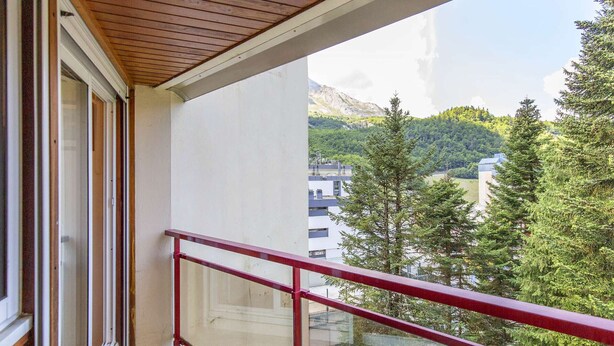 A balcony with a red railing, a wooden ceiling, and a view of a mountainous landscape with trees and buildings.