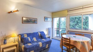 A cozy room with a blue sofa, a small table with a lamp, a dining table with chairs, and a large window with a view of trees.