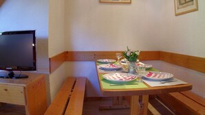 A dining area with a wooden table set for two, a television on a stand, and a framed picture on the wall.