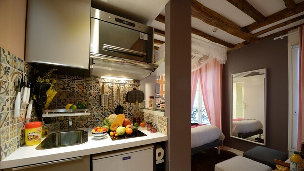 A compact kitchen with a built-in oven, a small refrigerator, and a sink. There is a dining area with a bed and a window with curtains. - E4GG So sweet (París)