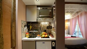 A compact kitchen with a refrigerator, microwave, and sink. A bed with a window and curtains is visible in the adjacent room. - E4GG So sweet (París)