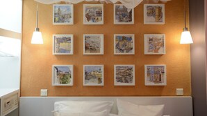 A hotel room with a bed, white pillows, and a wall decorated with framed cityscape paintings. - E4GG So sweet (París)