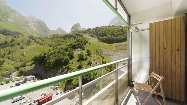 Un balcon donnant sur un paysage montagneux, une chaise en bois et un espace de stationnement en contrebas.