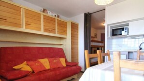 A cozy living area with a red sofa, wooden dining table, and chairs. A kitchen with a microwave is visible in the background. - Cozy Studio for 4 in Alpe d'Huez, Close to Slopes & Shops, Balcony, Pets Welcome (Huez)