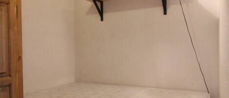 A small room with a single bed, a wooden door, and a wall-mounted shelf.