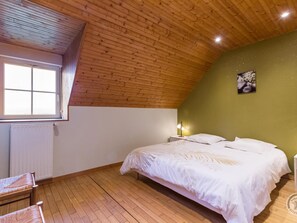 Quarto com teto de madeira inclinado, cama com roupa de cama branca, mesa de cabeceira com abajur e janela com persianas.