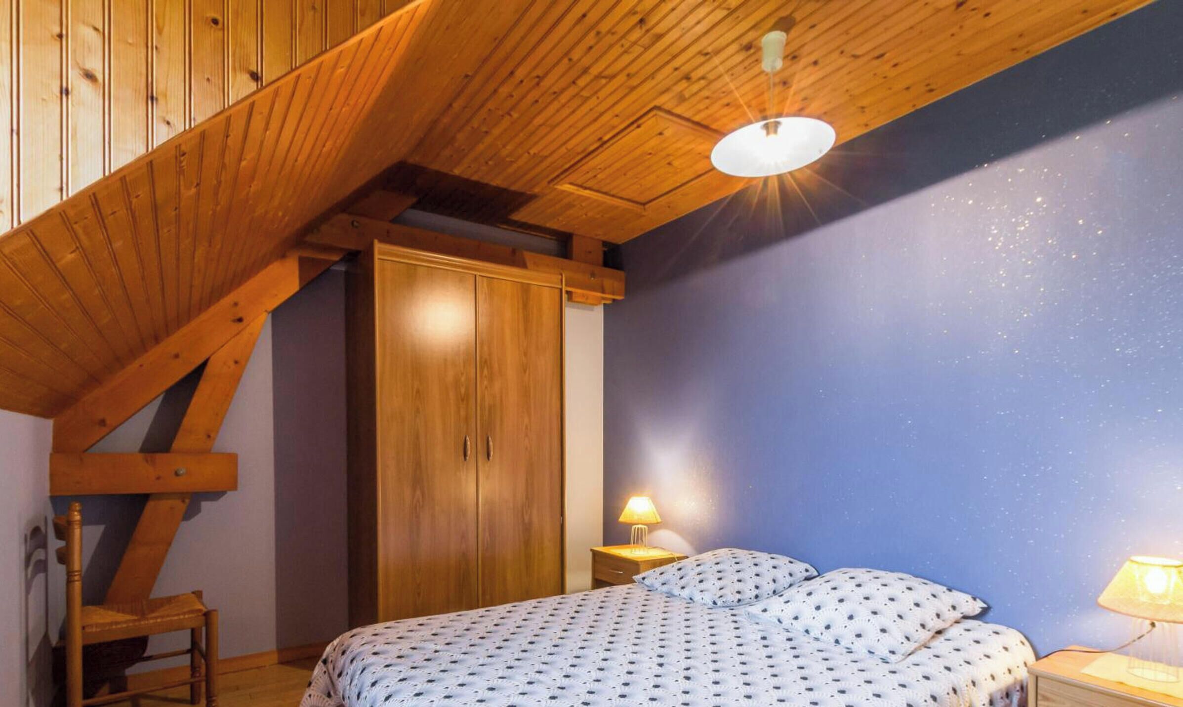 A bedroom with a sloped ceiling, wooden wardrobe, a bed with patterned bedding, and two bedside lamps.