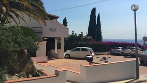 A modern building with a 'RENT' sign, a parking area with several cars, and a clear view of the sea in the background.