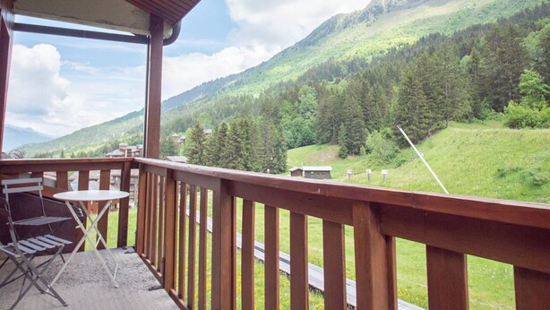 Un balcón con baranda de madera, una mesa plegable de metal y una silla, con vistas a un paisaje montañoso con vegetación y cielo despejado.