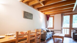 Una sala de estar acogedora con una mesa de comedor de madera, sillas, un sofá y una mesa baja. Se ve un ventanal con cortinas y vistas al exterior con vegetación.