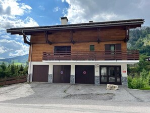 A two-story wooden building with a balcony, two garage doors, and a stone base. - Cozy apartment 7 sleeps, ski-in/ski-out, fireplace, parking (Le Grand-Bornand)
