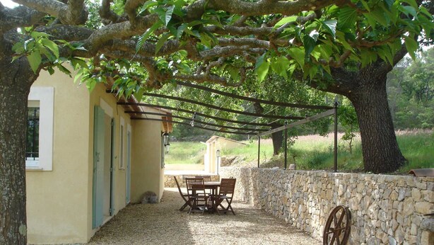 A covered patio with a table and chairs, a stone wall, and a tree providing shade.