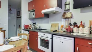 A compact kitchen with red cabinets, a white dishwasher, and a microwave. There is a wooden table with chairs, and various kitchen utensils and containers are visible. - Cozy 4-Person Apartment in Central Cauterets, Close to Ski Lift, Spa, and Amenities (Cauterets)