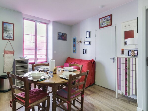 Comedor con mesa de madera dispuesta para cuatro personas, rodeada de sillas de madera con cojines rojos. Se ve una ventana con persianas rosadas, un armario blanco con cajones de colores y una puerta con espejo y lámpara.