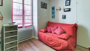 A room with a red sofa, a white radiator, a window with pink curtains, and several framed pictures on the wall. - Cozy 4-Person Apartment in Central Cauterets, Close to Ski Lift, Spa, and Amenities (Cauterets)
