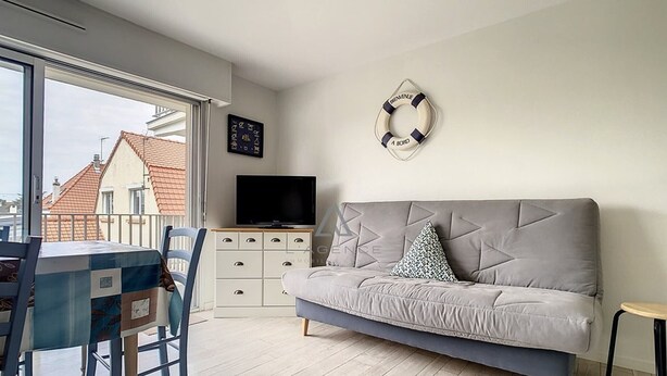 En moderne stue med en grå sofa, et fjernsyn på et hvidt skab, et spisebord med stole og udsigt til bygninger.